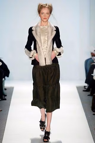 Runway model in a dark jacket, pale ruffled blouse and wide cropped bottoms.