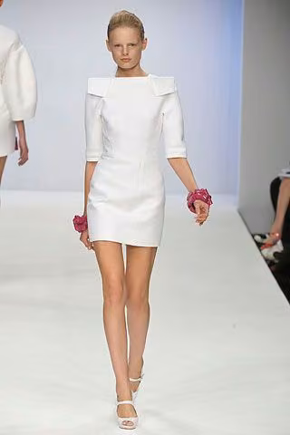 Runway model in a white minidress with a broad sculptural neckline.