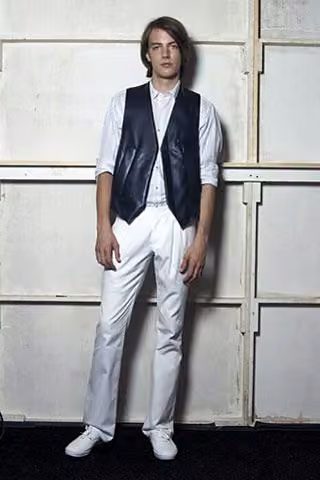 Model in a dark vest over a white shirt and white trousers.