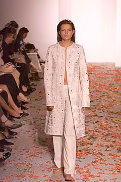 Runway model in a pale decorated long coat over pale trousers.