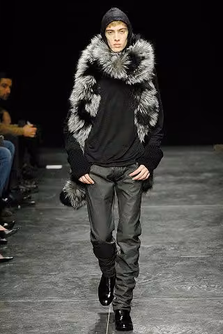 Runway model in a dark top with a large fur-trimmed hood and gray trousers.