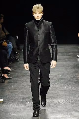 Runway model in a glossy black high-necked tailored jacket and trousers.