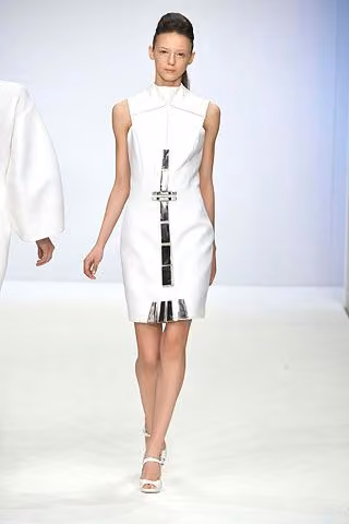 Runway model in a sleeveless white dress with a metallic vertical ornament.