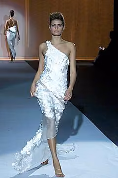 Runway model in a pale one-shoulder textured dress with a trailing hem.