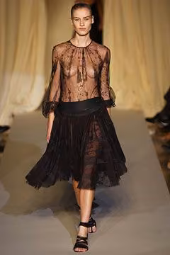Runway model in a sheer black lace blouse and full black skirt.