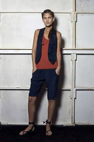 Model in a dark vest, rust-colored top and navy shorts.