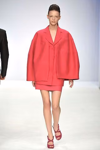 Runway model in a red voluminous jacket and short matching skirt.