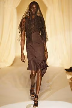 Runway model in a brown draped dress with a sheer upper layer.