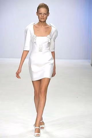 Runway model in a short white dress and cropped white outer layer.